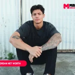 Dylan Jordan Net Worth article portrait of a man seated in black clothes against a white corrugated metal wall
