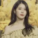 Lee Ji-Eun (IU) at a public event wearing a cream off-shoulder dress against a golden floral background