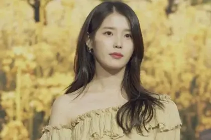 Lee Ji-Eun (IU) at a public event wearing a cream off-shoulder dress against a golden floral background