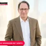 Andrew Weissmann portrait in brown tweed blazer, smiling indoors near sheer curtains