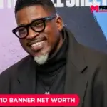 David Banner net worth portrait, smiling in glasses and black overcoat at event backdrop.
